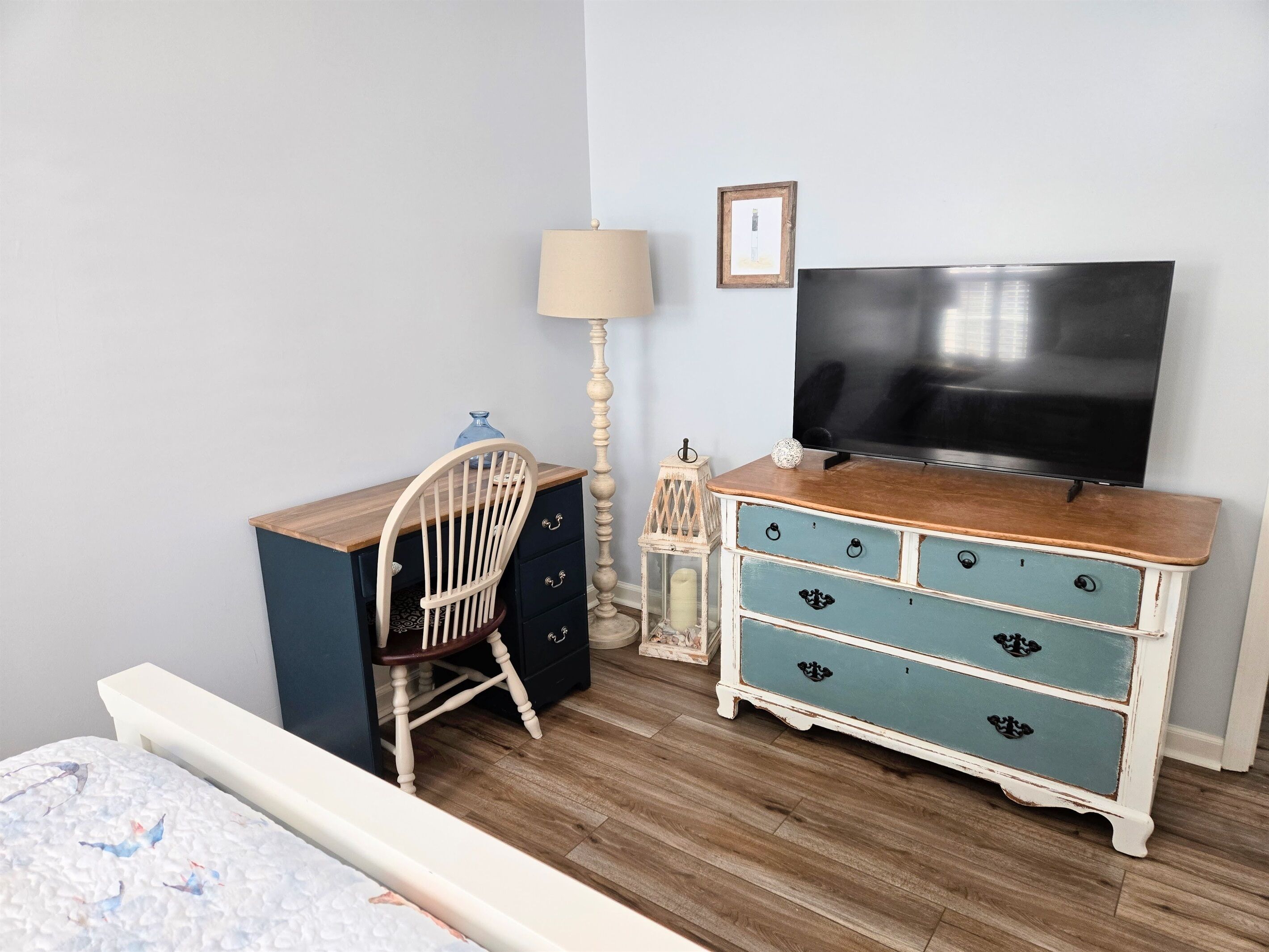 A bedroom with a wooden desk, a chair, a dresser, a television, and a lamp.
