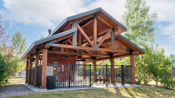 Situated in a serene landscaped area, this pavilion features a robust open framework with vertical beams and horizontal supports.