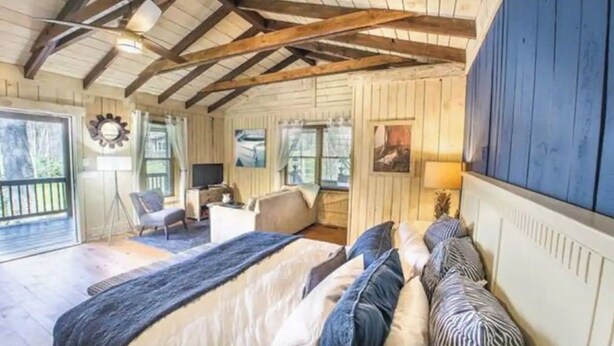 Un dormitorio acogedor con una cama grande, un escritorio y una silla. Hay una ventana con cortinas, una puerta que da a un balcón y un televisor montado en la pared.