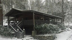 Una cabaña de madera con un balcón y unas escaleras que llevan a ella, rodeada de árboles cubiertos de nieve.