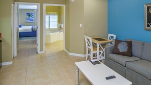 Un salon avec un canapé, une petite table et une chaise. Une porte donne accès à une chambre et à une salle de bain.