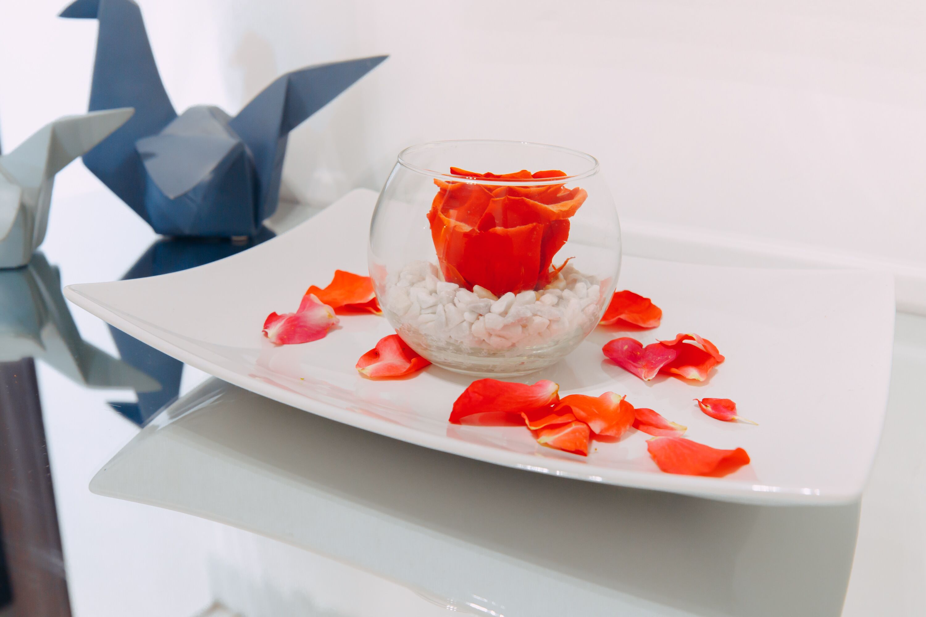 A glass bowl with a red rose and white pebbles on a white plate.
