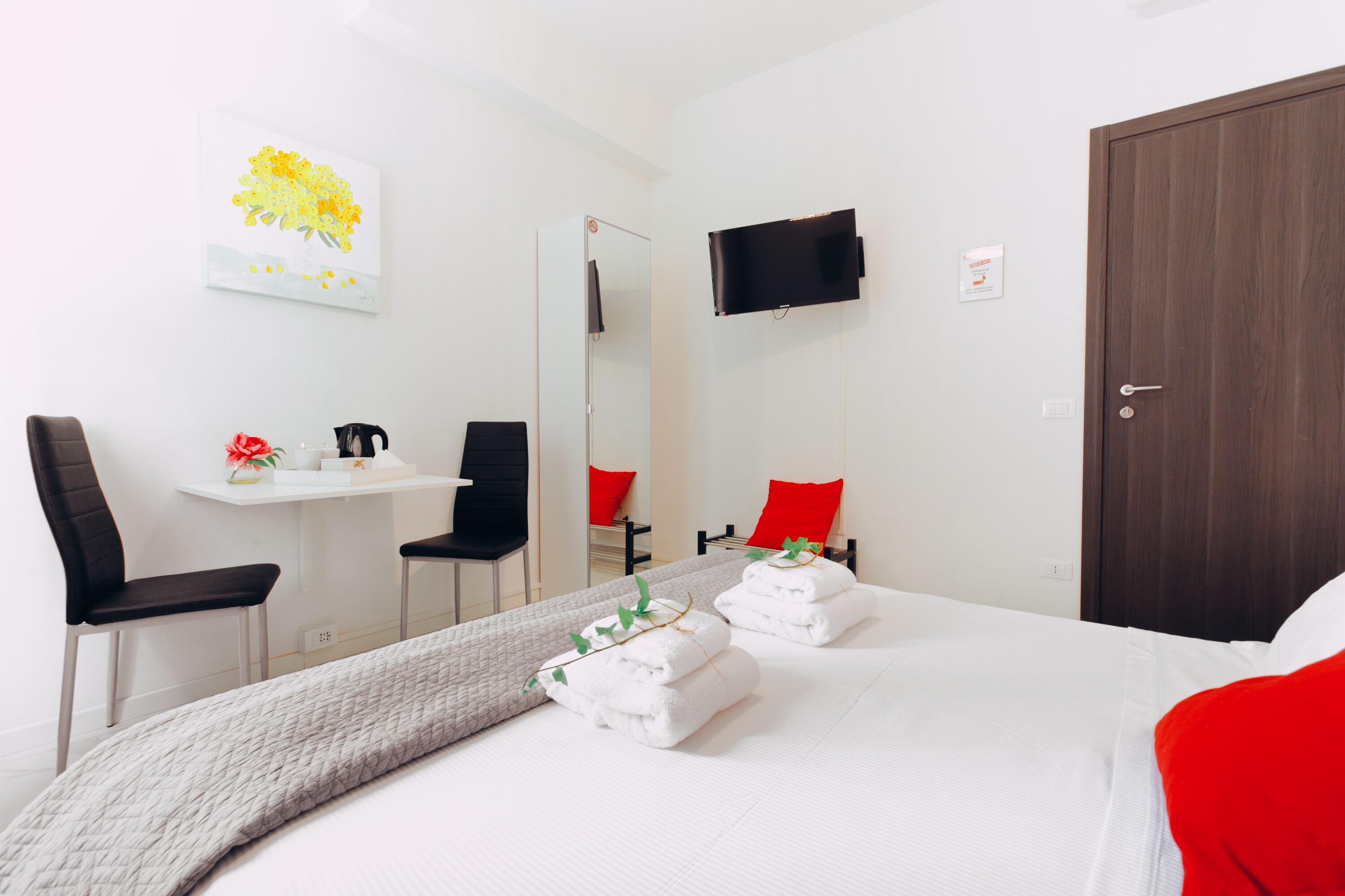 Una camera d'albergo con un letto, due sedie, un tavolino con un fiore rosso, una televisione montata a parete e una porta.