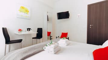 Una camera d'albergo con un letto, due sedie, un tavolino con un fiore rosso, una televisione montata a parete e una porta.