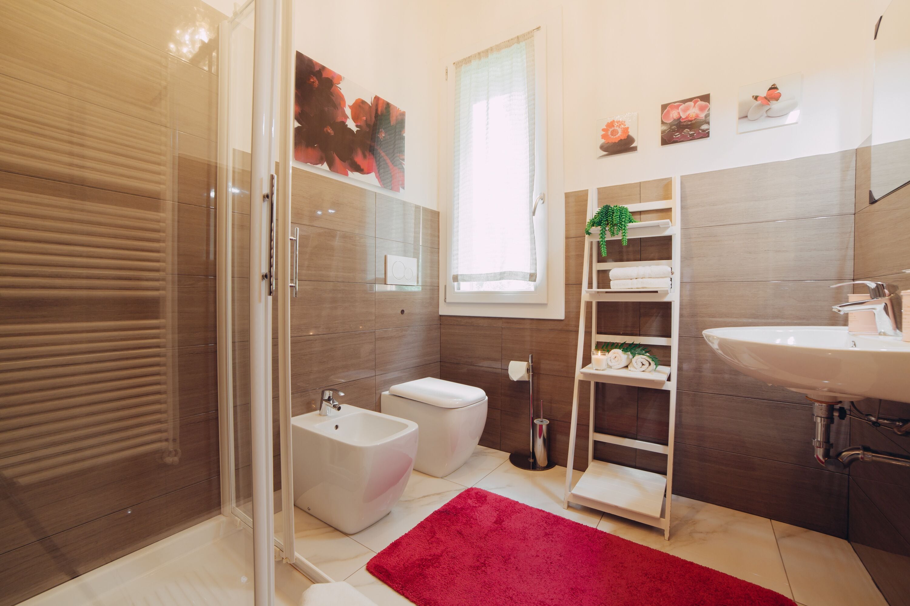 A modern bathroom with a glass shower enclosure, a bidet, a toilet, a sink, and a red rug.