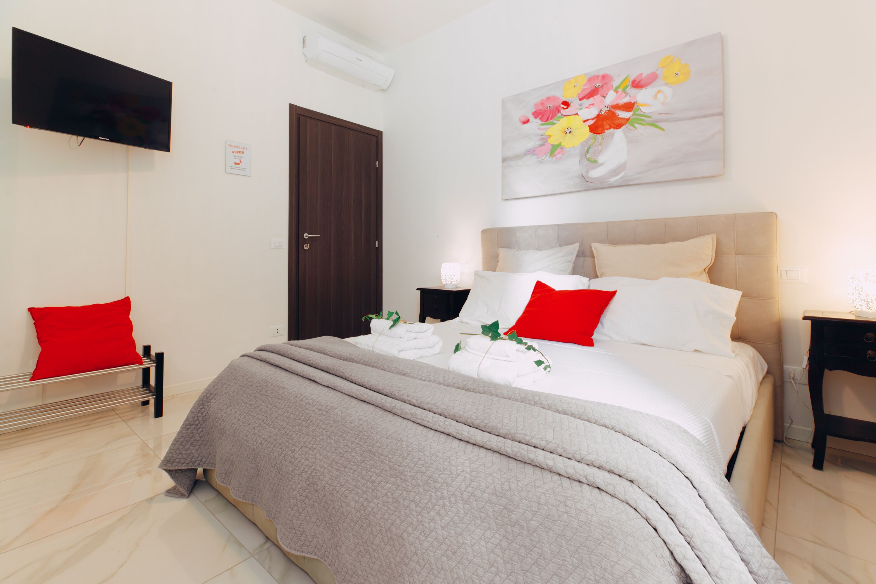 A neatly made bed with a grey blanket, a red pillow, and a white pillow. There is a flat-screen TV mounted on the wall, a wooden door, and a painting of flowers.