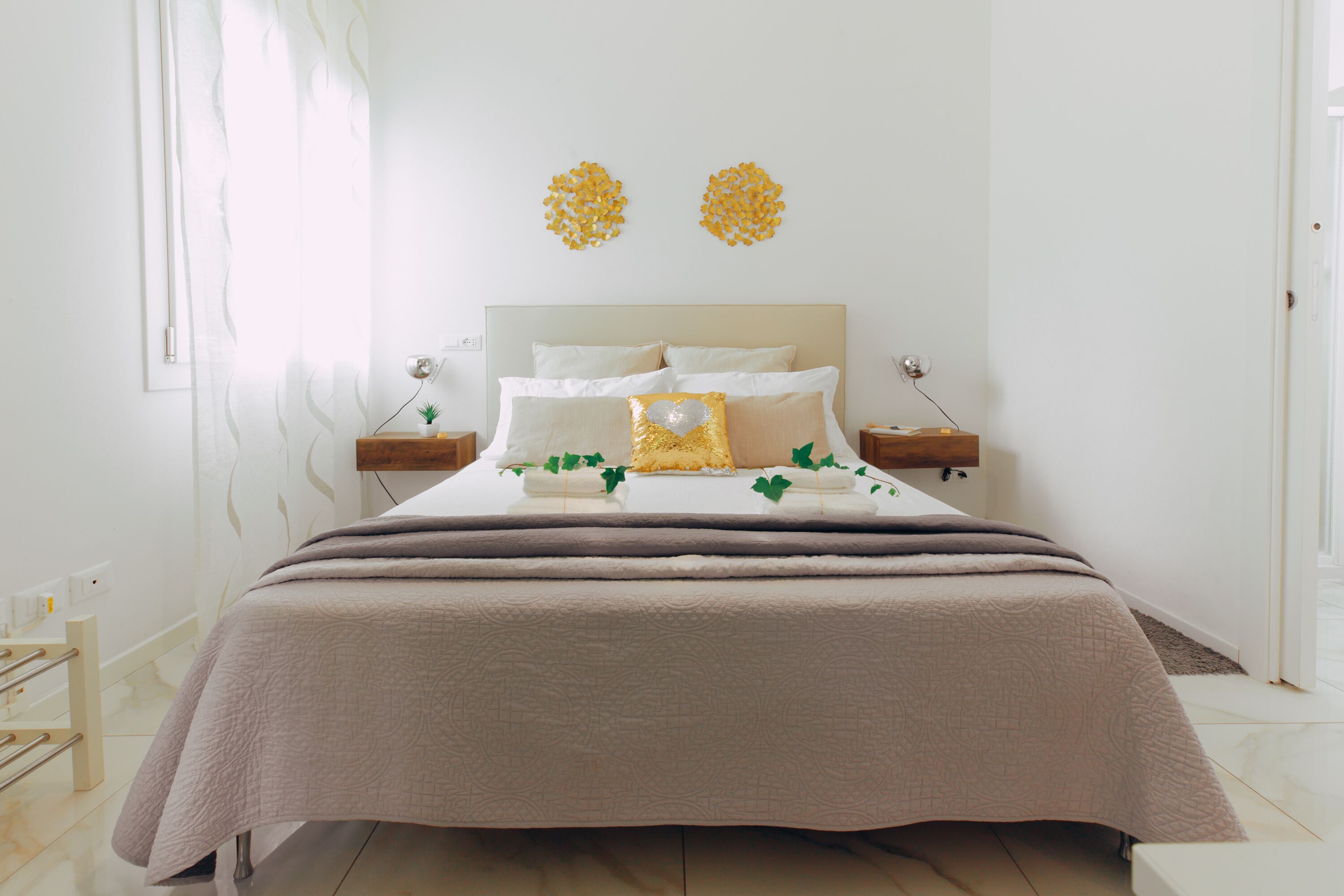 A neatly made bed with a beige bedspread and pillows, two bedside tables with lamps, and a window with sheer curtains.