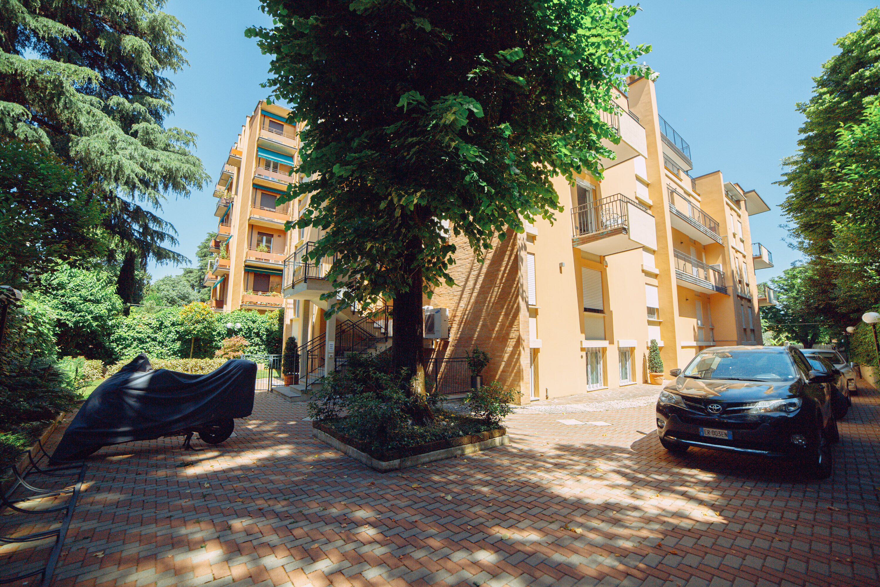 A multi-story residential building with balconies, a covered car, and a tree in the foreground.