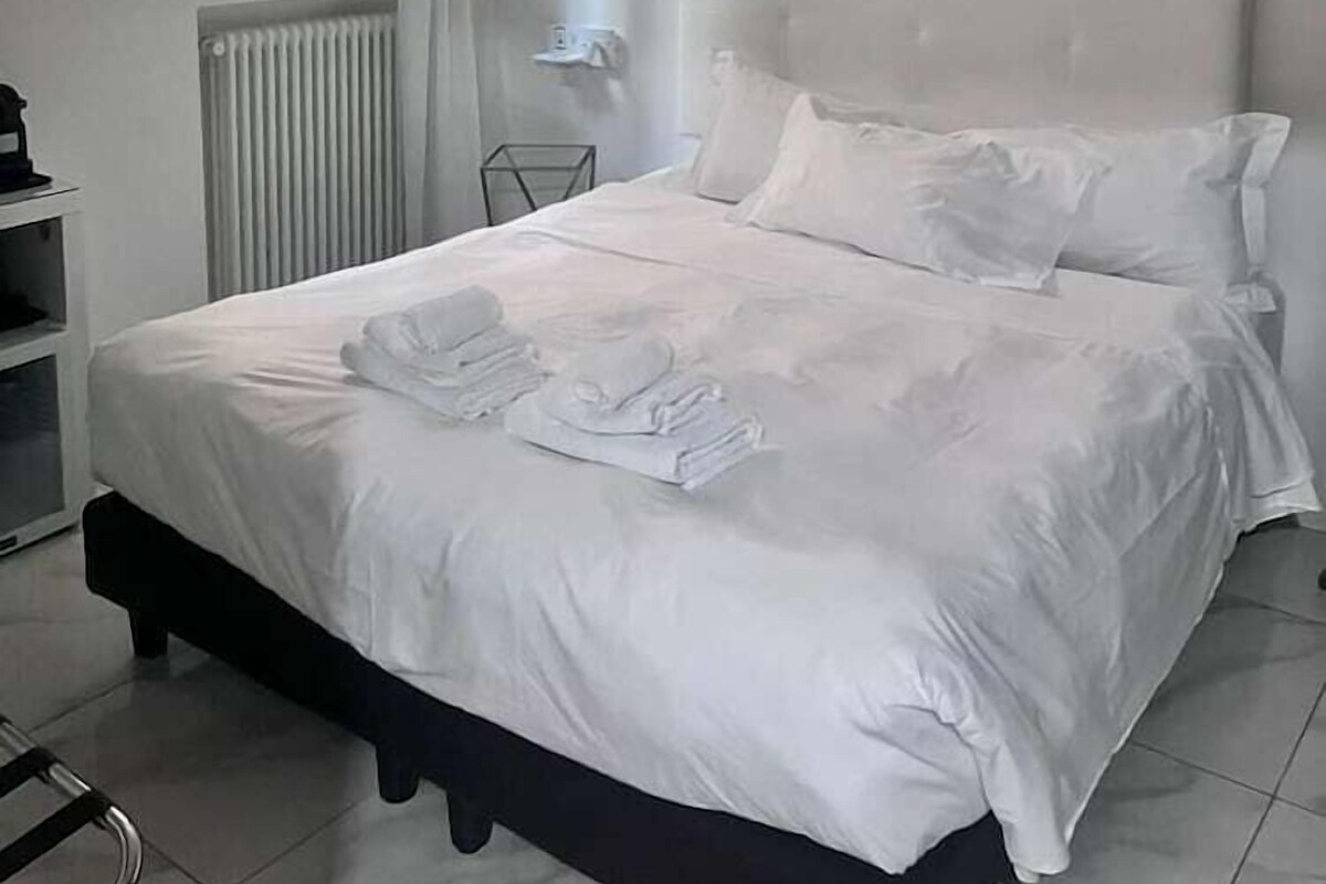 A neatly made bed with white linens and towels, a radiator, a window with curtains, and a wall-mounted artwork.