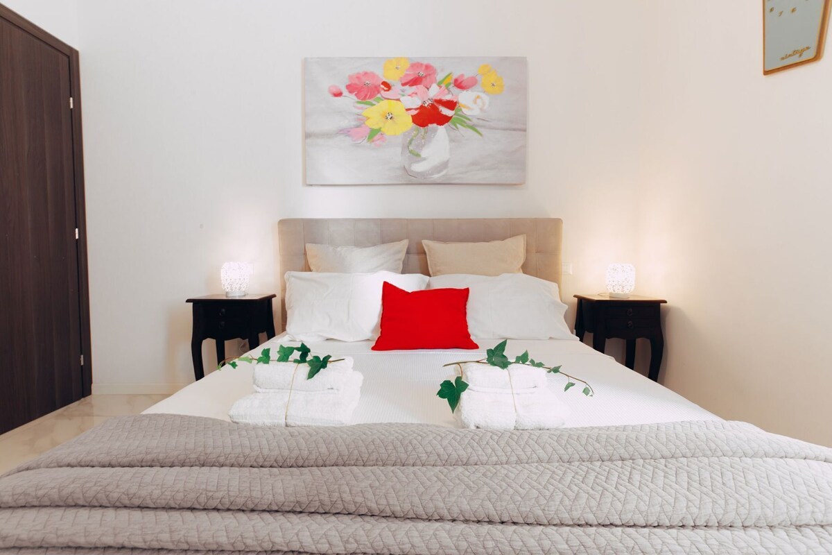 A neatly made bed with white bedding, a red pillow, and two bedside tables with lamps.