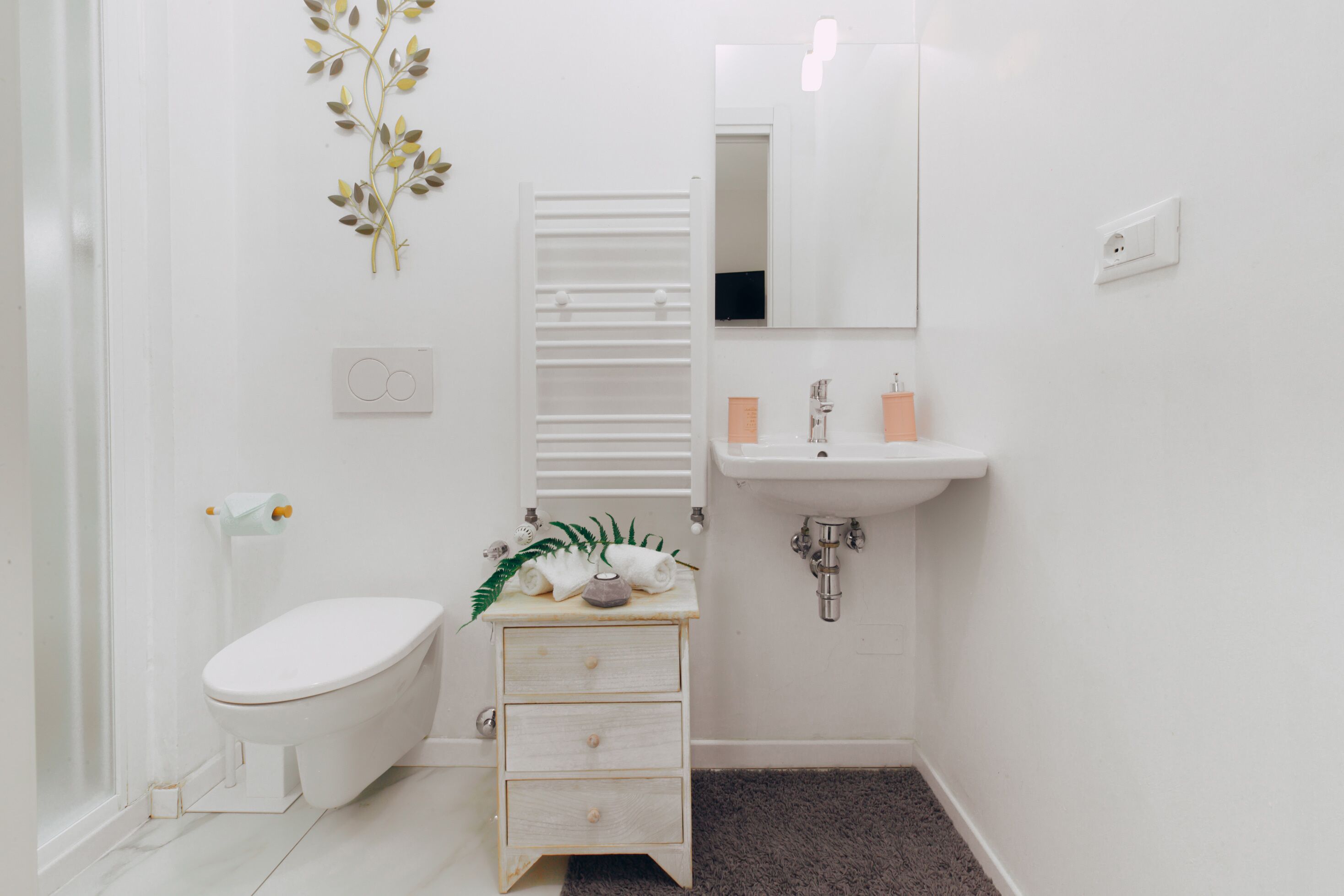 A small, white bathroom with a toilet, a sink, a mirror, and a small wooden cabinet.