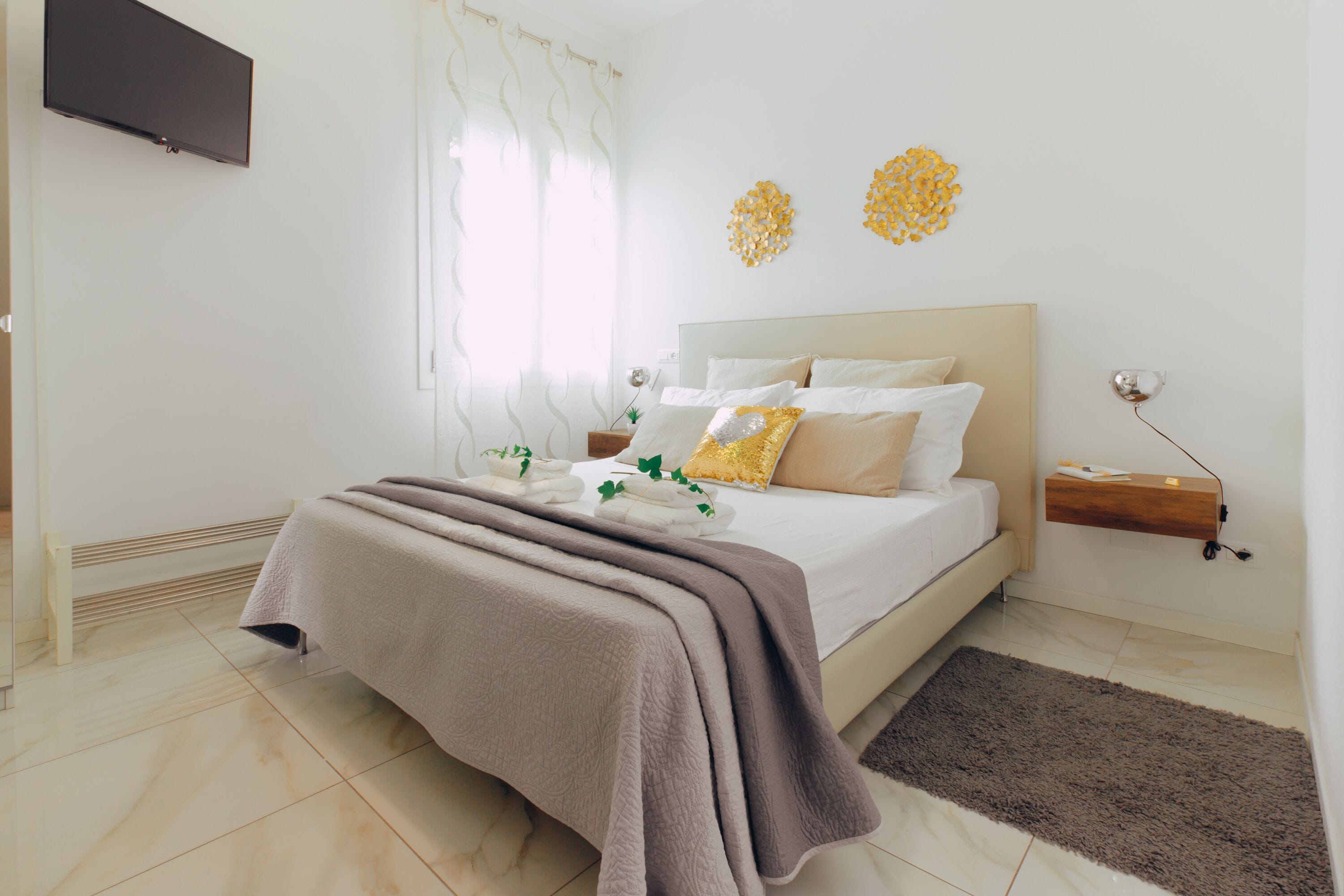 A bedroom with a bed, a bedside table, a TV mounted on the wall, and a window with curtains.