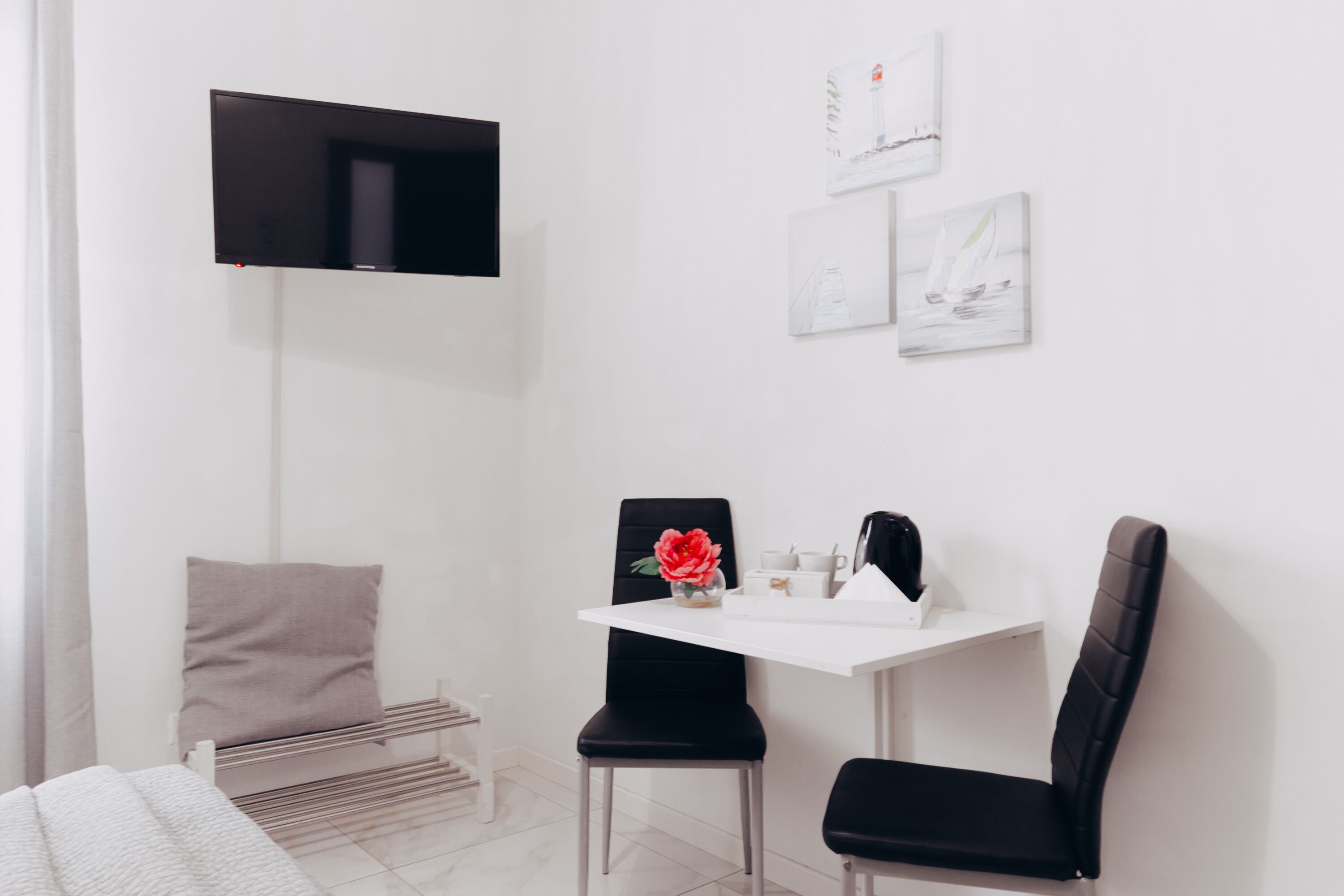 Una camera moderna e minimalista con un tavolo bianco, due sedie nere, una TV a schermo piatto montata a parete e una piccola panca.