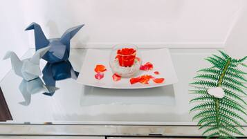 Una mesa con un cuenco de vidrio que contiene una rosa roja, rodeada de pétalos de rosa dispersos y un pájaro de origami azul y blanco decorativo.