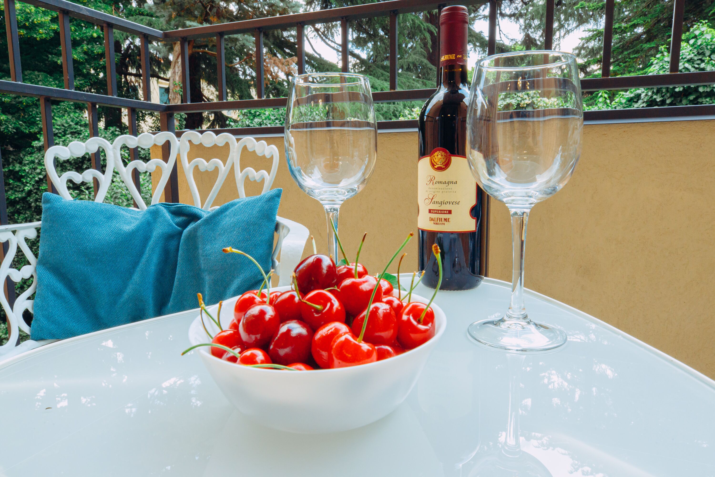 A table with a bowl of cherries, two wine glasses, and a bottle of wine on a balcony with a railing and greenery in the background.