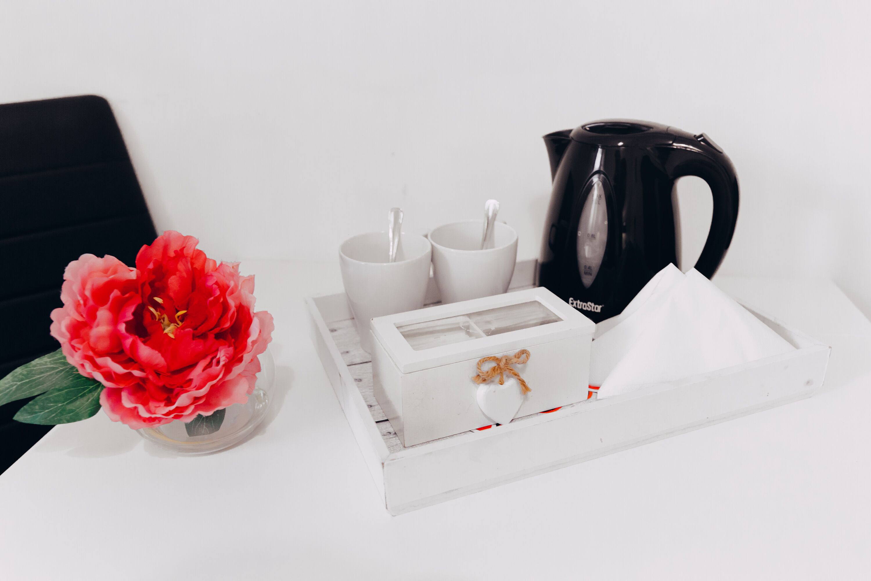 A white tray with a black coffee pot, two white cups, a white box with a heart-shaped lid, and a red flower in a clear vase.