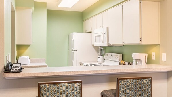 A kitchen with a refrigerator, microwave, stove, and coffee maker. There are two chairs with patterned upholstery.