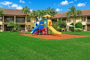 Ein Spielplatz mit Rutschen und Klettergerüsten, umgeben von einer gepflegten Rasenfläche und Palmen, mit einem Gebäude im Hintergrund.