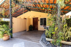 A covered outdoor area with a wooden floor, potted plants, and a door leading to a building.