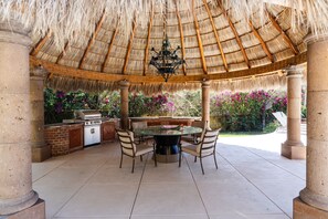 An outdoor patio with a thatched roof, a round table, chairs, a grill, and a brick wall with flowers.