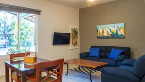 Ein Wohnzimmer mit einem dunkelblauen Sofa, einem Holztisch und einem Esstisch mit Stühlen. An der Wand hängt ein Gemälde, und es gibt einen Deckenventilator.