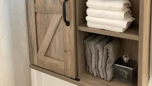 Ein Holzkabinet mit schwarzem Griff und Metallbeschlägen, das gefaltete weiße und graue Handtücher sowie eine kleine schwarze Schachtel enthält.