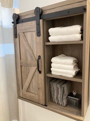Ein Holzkabinet mit schwarzem Griff und Metallbeschlägen, das gefaltete weiße und graue Handtücher sowie eine kleine schwarze Schachtel enthält.