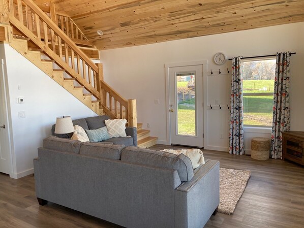 Ein Wohnzimmer mit grauem Sofa, einer Holztreppe und einer Tür, die nach draußen führt.
