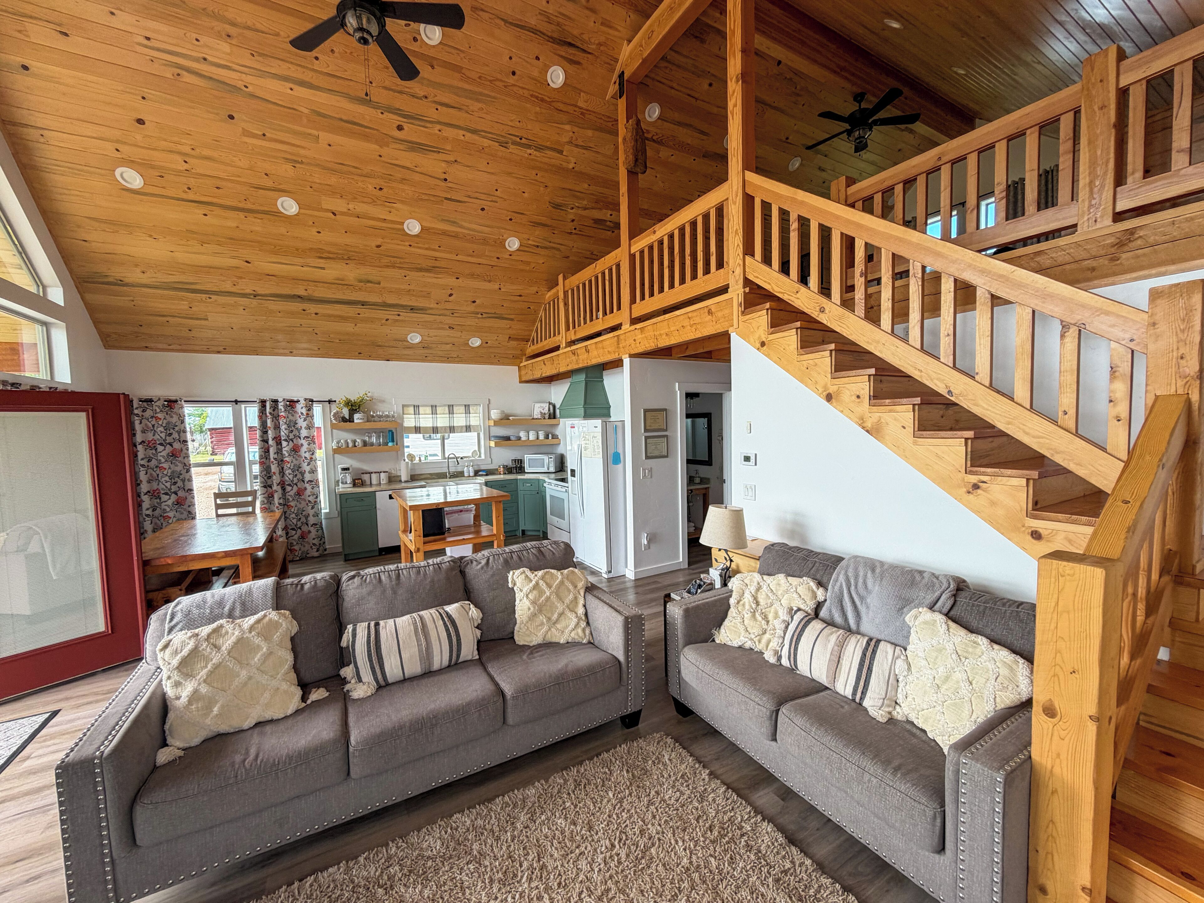 A spacious living area with a wooden staircase, a grey sofa set, and a kitchen in the background.