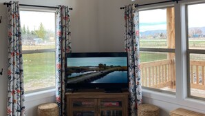 Ein Wohnzimmer mit einem großen Flachbildfernseher auf einem Holzboden, umrahmt von Fenstern mit blumigen Vorhängen, einer Holzbalkendecke und Blick auf eine Landschaft.