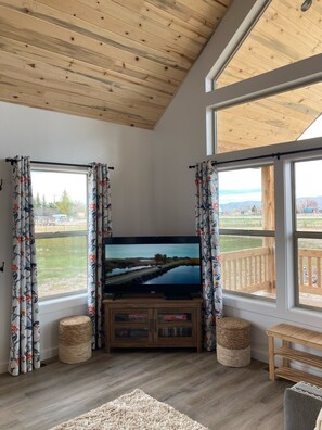 Ein Wohnzimmer mit einem großen Flachbildfernseher auf einem Holzboden, umrahmt von Fenstern mit blumigen Vorhängen, einer Holzbalkendecke und Blick auf eine Landschaft.