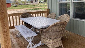 Eine überdachte Veranda mit Holztisch und Korbstühlen, mit Blick auf eine ländliche Landschaft mit Scheunen und Feldern.