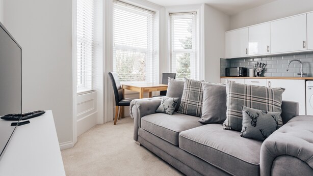 Ein modernes Wohnzimmer mit grauem Sofa, einem Esstisch und einer Küchenzeile mit weißen Schränken und einer Mikrowelle.