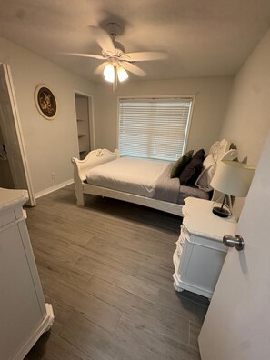 A bedroom with a bed, a ceiling fan, a window with blinds, and a bedside table with a lamp.