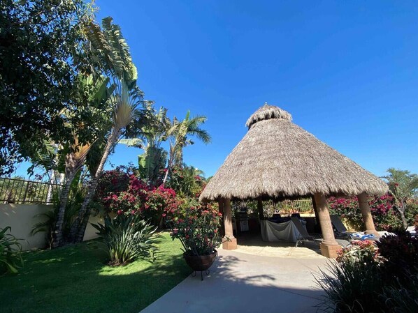 A thatched-roof structure with a paved walkway leading to it, surrounded by lush greenery and flowering plants.
