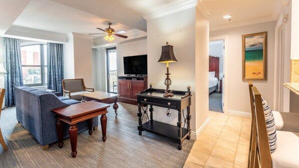 A hotel room with a sofa, a wooden coffee table, a desk with a lamp, a television, and a painting on the wall.