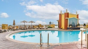 A pool area with lounge chairs and umbrellas, a fountain, and a building with a tiled facade.