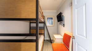 Une chambre avec des lits superposés, un bureau, une chaise avec un coussin orange, un téléviseur fixé au mur et une porte.