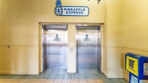 Une paire de portes d’ascenseur ornées du logo de « Pineapple Express », un sol carrelé et un distributeur automatique situé à droite.