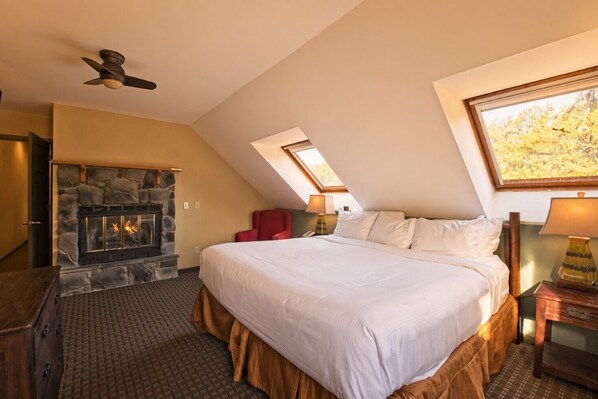 A bedroom with a stone fireplace, a bed with white linens, a red chair, and a ceiling fan.