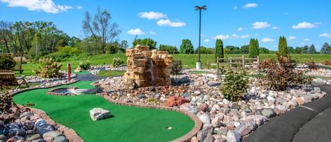 Un campo de mini golf con una superficie de césped verde, una formación rocosa como obstáculo y una valla de madera al fondo.