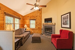 Een gezellig interieur van een chalet met een open haard, houten wanden, een plafondventilator en een rode fauteuil.