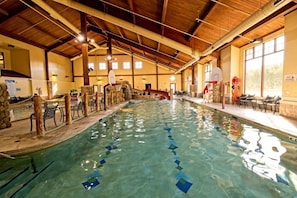 An indoor swimming pool with a wooden ceiling, a diving board, and a seating area with tables and chairs.