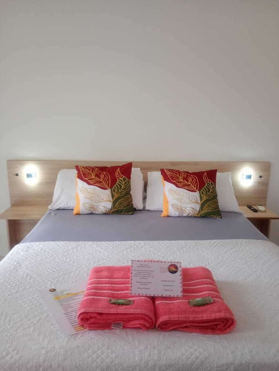 Una cama bien hecha con una toalla doblada y una tarjeta, dos almohadas con motivos de hojas, y lámparas de noche.