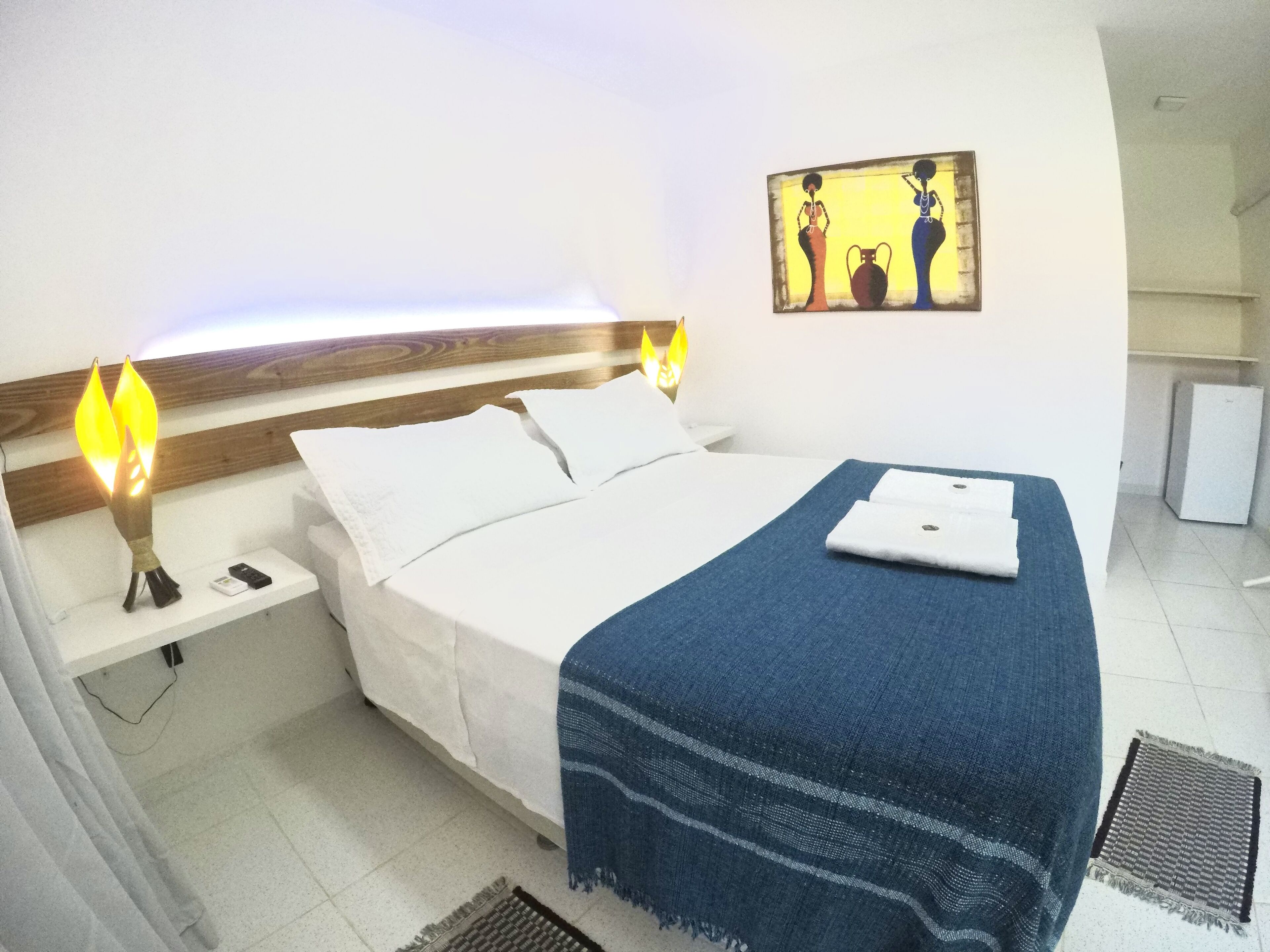 A hotel room with a bed, bedside lamps, a painting, and a small refrigerator.