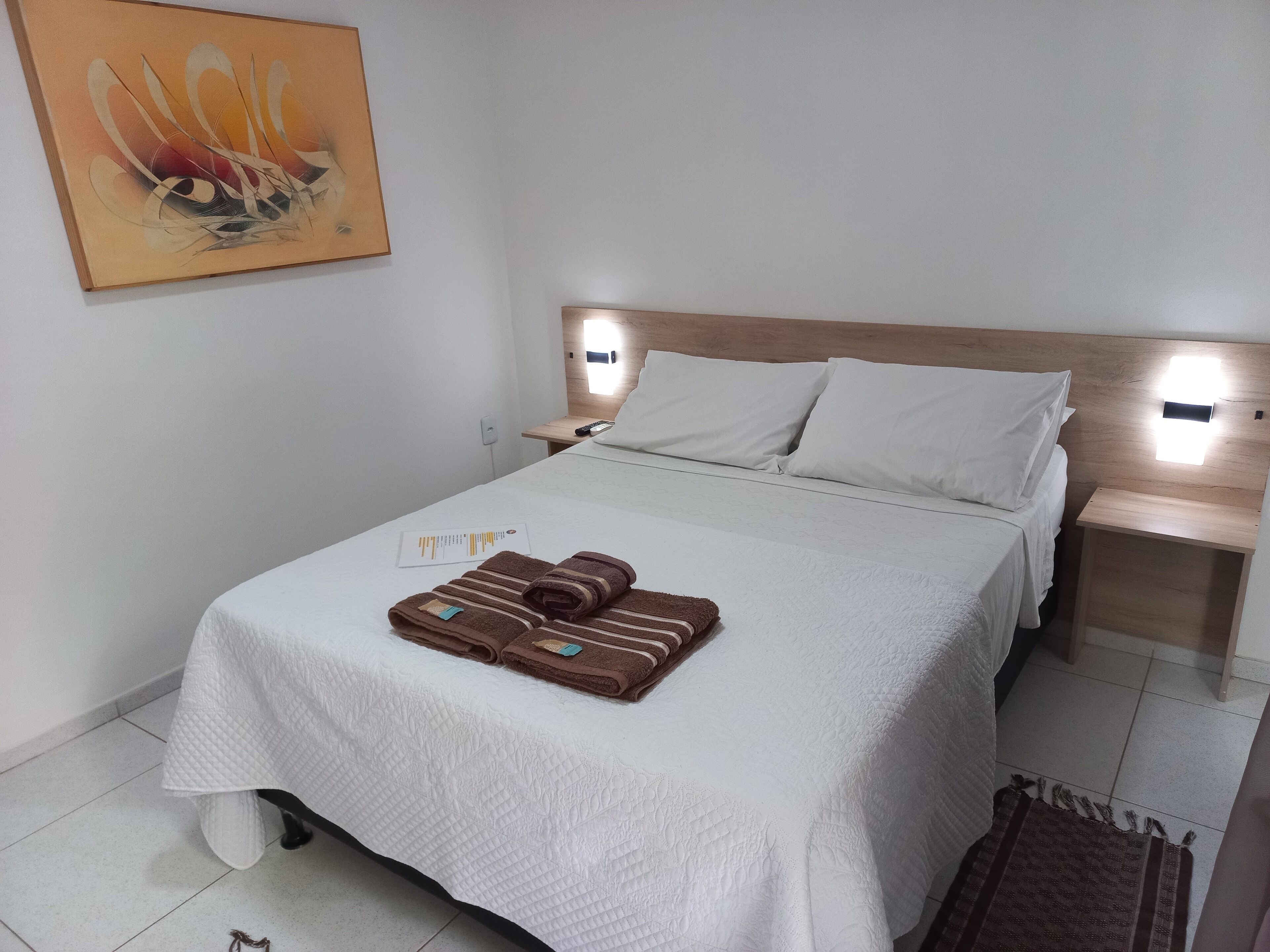 A neatly made bed with white bedding and brown towels, a wooden headboard, and a painting on the wall.