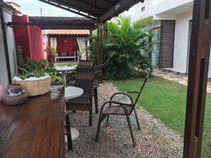 Ein sonniger Außenbereich mit einer Terrasse, Tisch und Stühlen, umgeben von Grünpflanzen und einem kleinen Rasen.