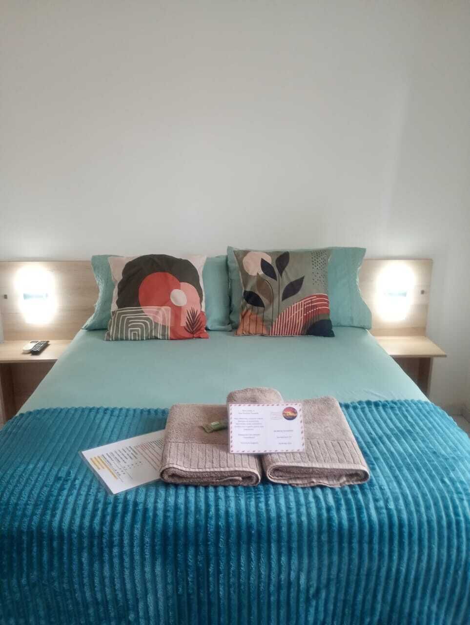 Una cama con colcha verde azulada, dos almohadas con diseños abstractos, un control remoto y una toalla doblada con una etiqueta sobre una mesita de noche de madera.