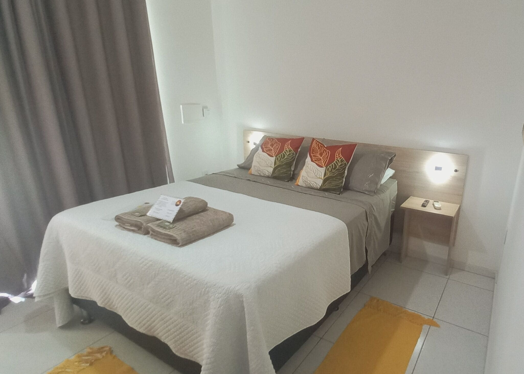 A neatly made bed with a white bedspread and two decorative pillows, a bedside table with a remote control, and a small rug on the floor.