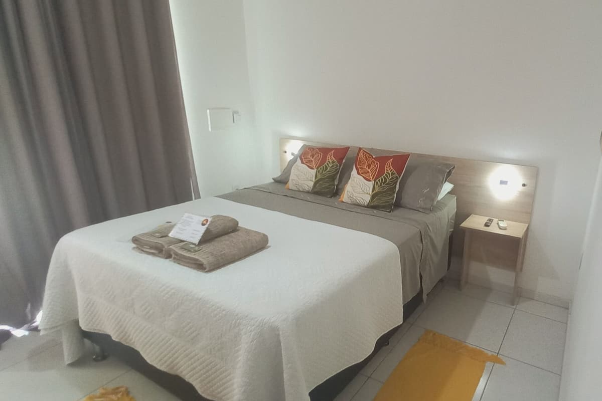 A neatly made bed with a white bedspread and two decorative pillows, a bedside table with a remote control, and a small rug on the floor.